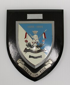 Black wooden shield with shield-shaped emblem in blue and white with a silver border. silver ribbon at bottom has a Latin motto.