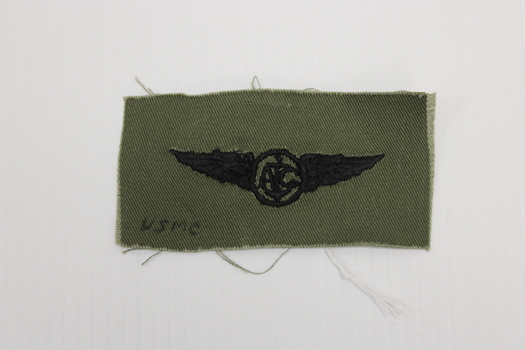 Rectangle Khaki coloured textile badge with USMC insignia.  Black embroidery of a circle with wings each side. the letter A and C with an anchor between.