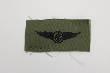 Rectangle Khaki coloured textile badge with USMC insignia.  Black embroidery of a circle with wings each side. the letter A and C with an anchor between.
