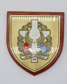 Wooden shield shaped plaque with gold shield shaped interior. A White cross with 50 in gold in centre, two hands clasped in hand shake with Vietnam Flag on left and Australian Flag on right.  Black and gold wreath surrounds image.