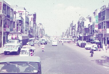 View down centre of main road, buildings down sides with parked and traveling vehicles.
