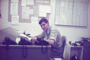 Sgt Ray Penny at his desk, 3 phones and chart on wall.