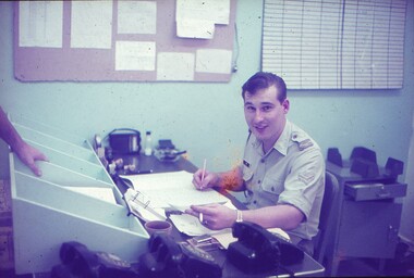 Cpl Peter Sims at his desk working with cigarette in left hand and pen in right.