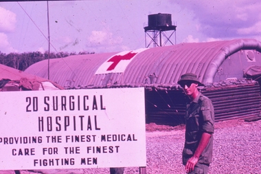 Soldier standing by sign for 20 Surgical Hospital with medical red cross on building in background.