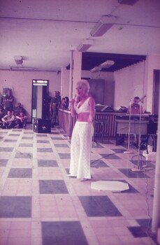 Female singer in white bikini top and white long pants.