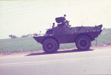 Armoured vehicle on side of road, with 2 personnel sitting at hatches.