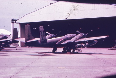 An aircraft parked outside a hangar. A bomb attached to the underside of the wing.