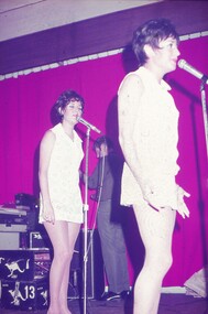 Two female entertainers in white dresses performing at microphones.