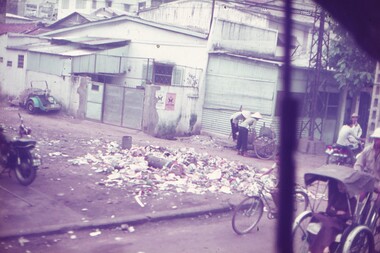 Dirt road, buildings and people on bikes and rickshaws going about their daily life.