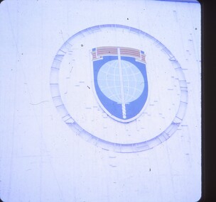 Shield shaped badge with scroll at top, circle in middle and sword running vertically from bottom to top.