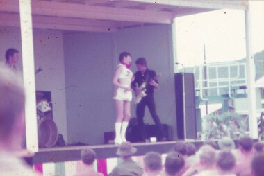 Female entertainer in white shorts top and knee high boots on stage with guitarist dressed in black, in the background.