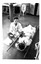 Black and white photo of an injured person on a stretcher. Another person is seated on a mat fanning the patient. 