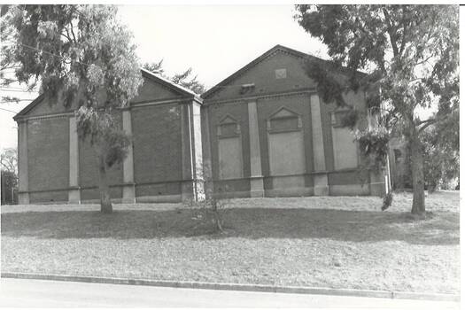 Black and white photograph of double fronted brick building with relief concrete columns, the windows and door of the right hand side filled in.