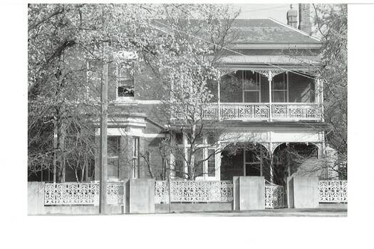 B/W photograph, double-story brick dwelling, bay window, verandahs both floors with iron lace. Iron lace fence, brick pillars, trees in front yard.