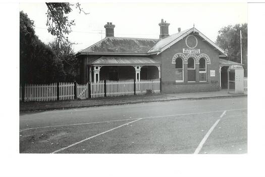 L-fronted polychrome brick building, slate roof, picket fence, covered verandah left side, three arched windows right side, telephone booth to the right.