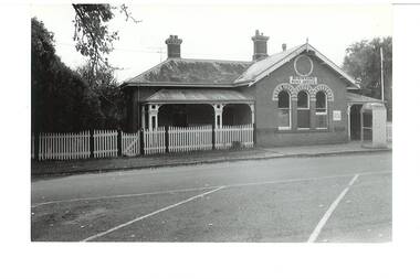L-fronted polychrome brick building, slate roof, picket fence, covered verandah left side, three arched windows right side, telephone booth to the right.