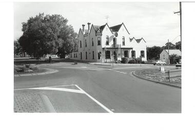 B/W photo of two-story, gabled hotel, brick painted white, viewed across intersection, large chinese elm tree to left