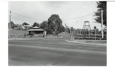 Original B/W photograph of streetscape of De Soza park frontage to Warrenheip St., Buninyong, with poppet head, rotunda and Chilean Wheel sculpture, former post office in background. Large eucalypt tree present behind rotunda, large cypress tree to north of park.