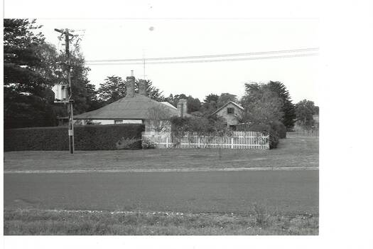 Side view of weatherboard house and brick outbuilding across road, tall hedge and white picket fence. Telegraph pole and sub-station.