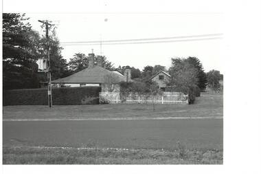 Side view of weatherboard house and brick outbuilding across road, tall hedge and white picket fence. Telegraph pole and sub-station.
