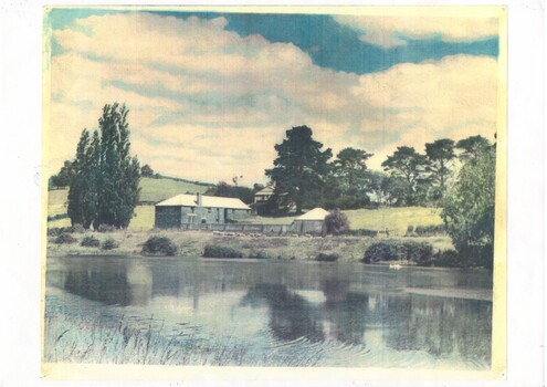 Hand-tinted photo across reservoir, fields rising behind old bluestone brewery and out-building, house obscured by trees behind.