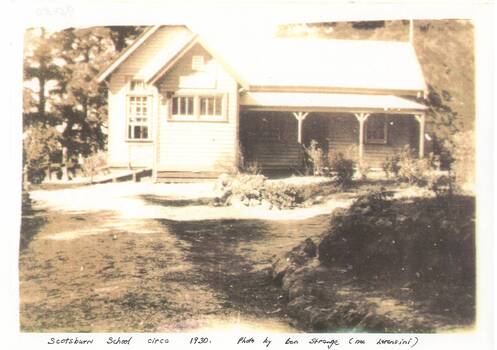 Sepia photo of school building across yard, gable roof, verandah across front right, tall trees behind