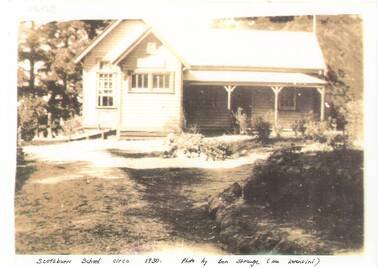 Sepia photo of school building across yard, gable roof, verandah across front right, tall trees behind