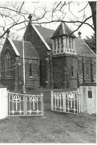 B/w photo, 3/4 view through white wrought iron gates,  bluestone chiourch, gables, low square bell tower.