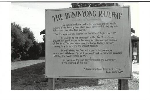 B/W photo, rectangular sign, on grass, trees surrounding, part of veranday behind.