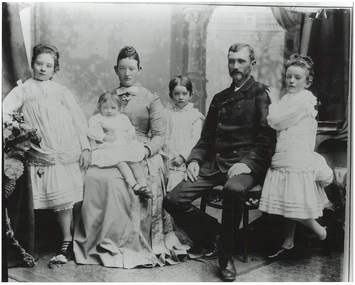 B/W studio portrait of family, parents (seated) and five children, all well dressed, children in white, standing except youngest son in mother's arms