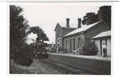 Bluestone station building on right, two story, three chimneys. Flower beds and man on platform, Steam train pulling in from left.