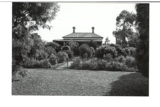 View from nature strip through heavily planted garden obscuring most of house, veranda just visible, high iron roof and two chimneys.