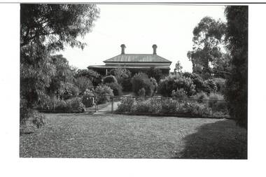 View from nature strip through heavily planted garden obscuring most of house, veranda just visible, high iron roof and two chimneys.