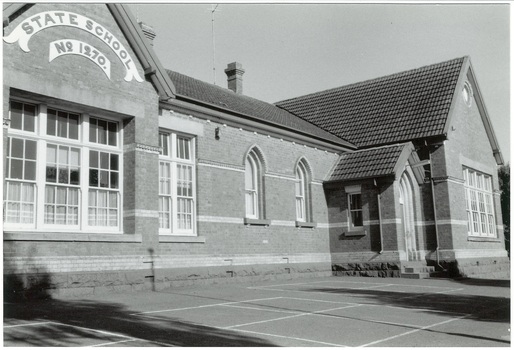 Polychrome brick school building, single story, tile pitched roof, original gothic style arched windows and door, and square replacement windows. Asphalt foreground.