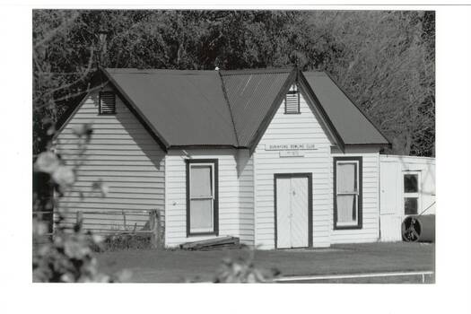 White painted wooden building, gabled tin roof, two tall windows, plank double doors. Greens roller on right.