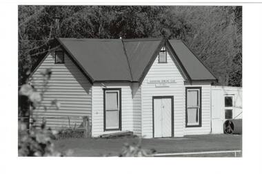 White painted wooden building, gabled tin roof, two tall windows, plank double doors. Greens roller on right.