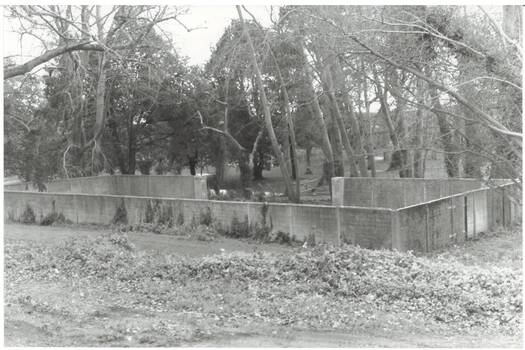 Tall concrete wall enclosing rectangular space, 1/3 gap in far wall, tall trees in Gardens behind