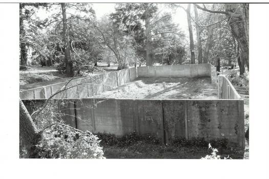 Looking over tall concrete walls into rectangular space, bare earth inside, surrounded by tall trees 