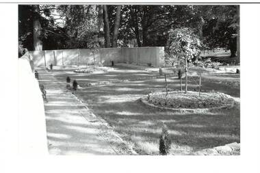 Inside walled garden, concrete walls, two round garden beds, new plantings of trees, bluestone edgings. Trees beyond walls.