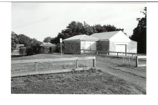 Sandstone building, low pitched iron roof, with Brick extension to right, iron roof. Small brick building and trees at rear