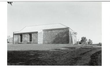Low brick building, small skillion extension with two tall windows to left. Rear wall of sandstone building at right.