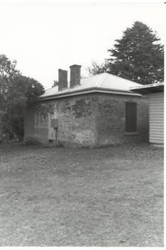 Small brick building, door back wall, window side wall, two chimneys, tall trees behind.