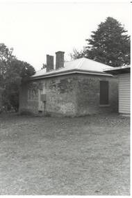 Small brick building, door back wall, window side wall, two chimneys, tall trees behind.