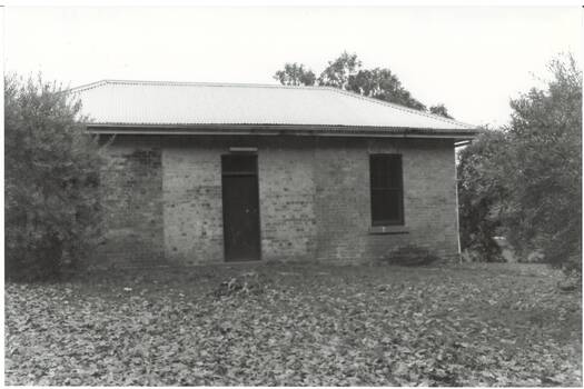 Small brick building, tall narrow windows flank central doorway, low pitched iron roof, trees surrounding