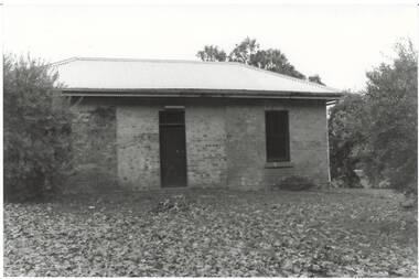 Small brick building, tall narrow windows flank central doorway, low pitched iron roof, trees surrounding