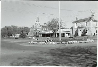 Streetscape of Learmonth St., Buninyong, looking over roundabout to Town Hall and businesses. Trees in the grounds of the Uniting Church are seen in the background. Buildings are the fire station, council offices, Town Hall, Whykes Butchers, D. Brown electrician, Buninyong Cake Shop and former bank, now a residence.