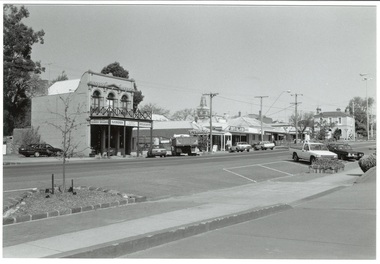 Streetscape across paved road, cars and trucks parked, double story building with verandah centre, then single story shops, and two story bank across roundabout.