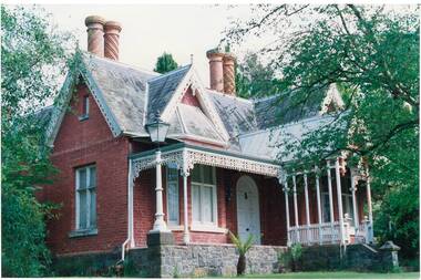 Colour photo of "Clifton Villa", cnr Eyre and Winter Sts, Buninyong. Built c.1860 for Mr Newman. The red brick building has bluestone foundations, cast iron lacework verandah trim, high ornate gables, slate roof and unusual round brick chimneys with spiral brick decorations.