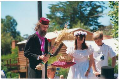 Bearded man in frock coat and fez, holding sheaf of wheat, girl in white dress holding red cusion and crown, next to boy on t-shirt.