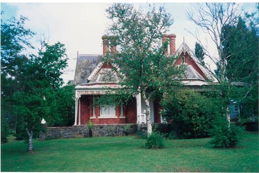 Colour photo of "Clifton Villa", cnr Eyre and Winter Sts, Buninyong. The red brick building has bluestone foundations, cast iron lacework verandah trim, high ornate gables, slate roof and unusual round brick chimneys with spiral brick decorations.